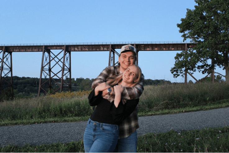 The couple embracing on a path beneath a railroad trestle at dusk.